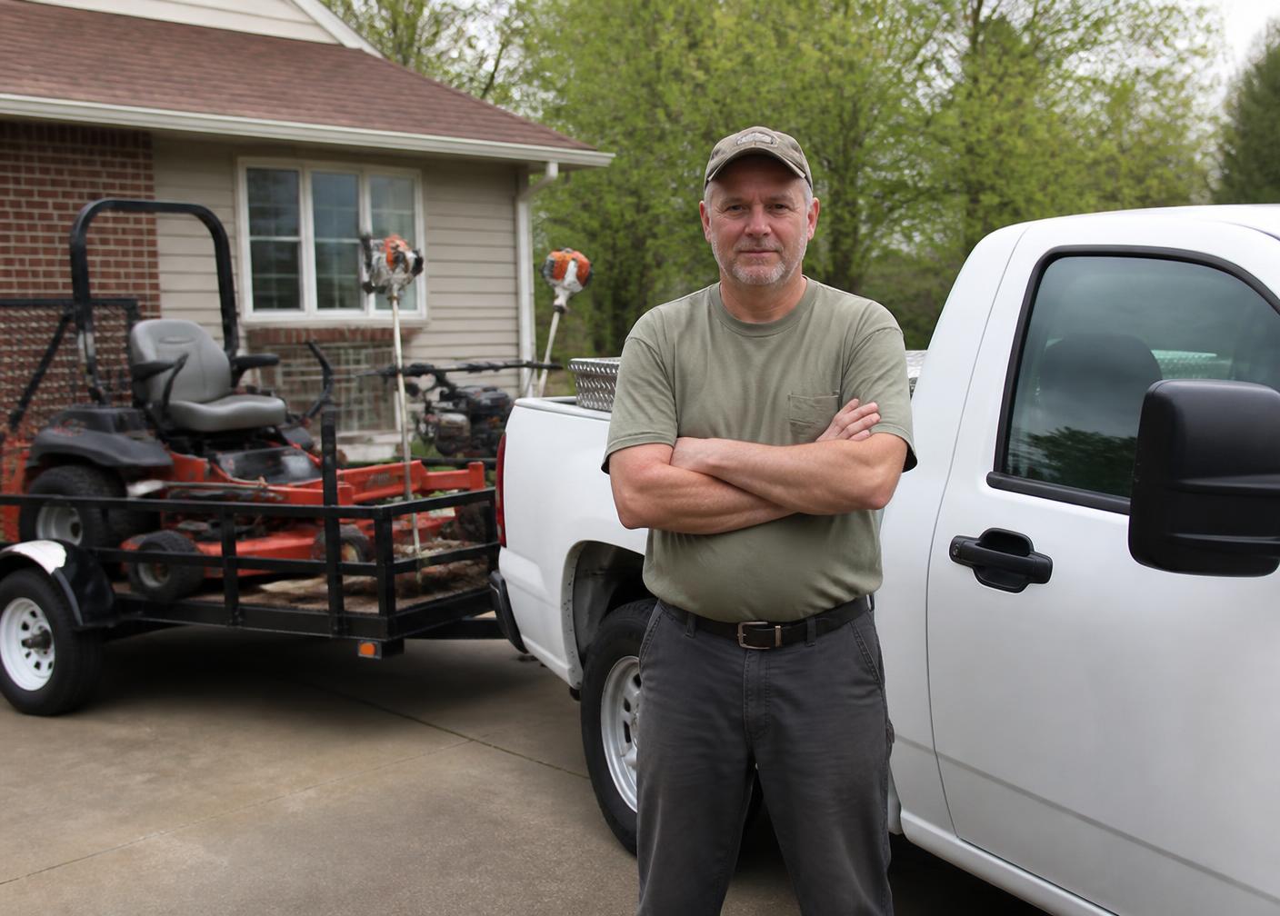 A lawn-care business owner standing beside his work truck and equipment trailer